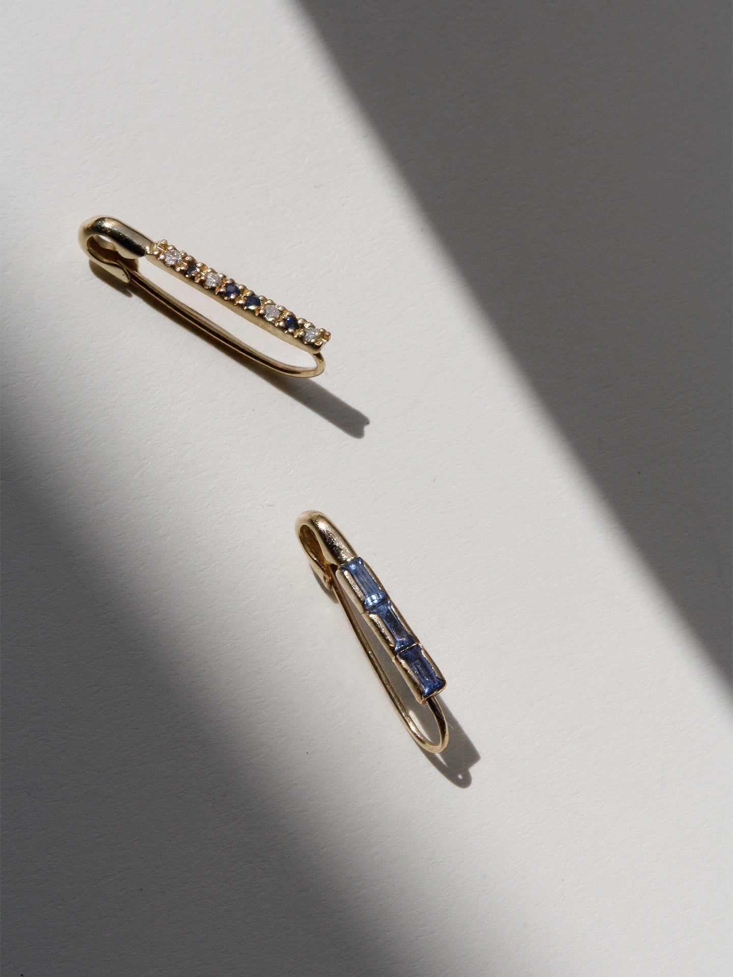 Flat lay of 14kt Yellow Gold Mini Sapphire Baguette Safety Pin Earring pictured with the new Mini Diamond & Sapphire Safety Pin.
