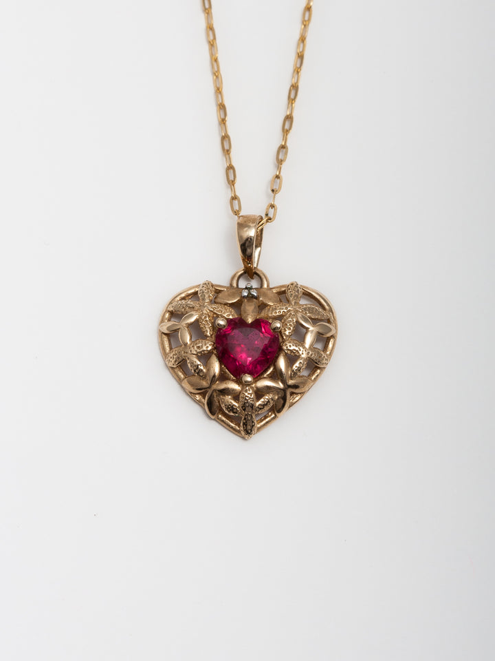 Close up picture of 14Kt Yellow Gold Heart Pendant with CZ Ruby Heart pictured on chain. Light grey background. 