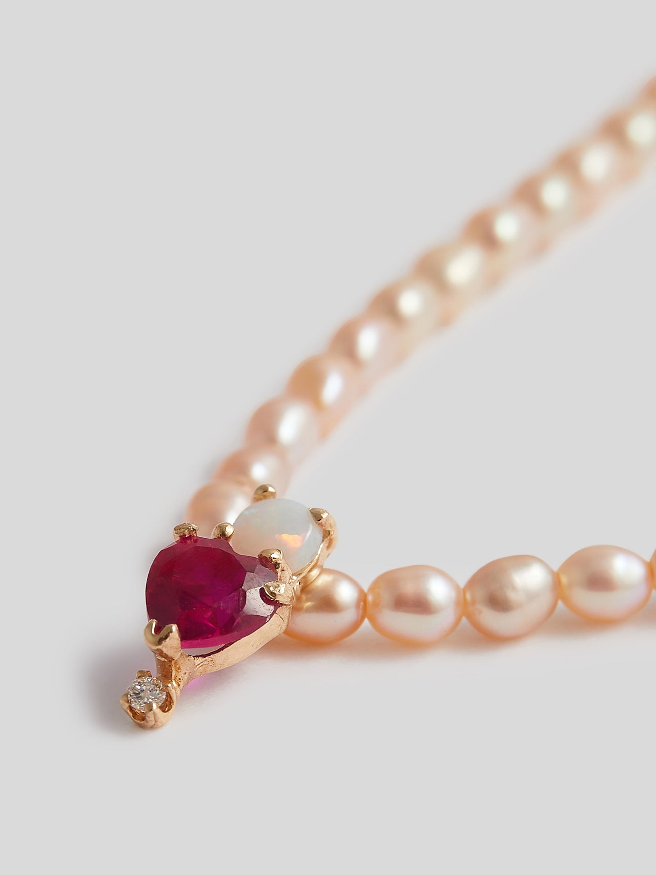 Close up product shot of a pink freshwater pearl necklace with a pendant that is a red heart gem with a small round opal gem stacked on top and diamond at bottom of pendant. Background is white. 