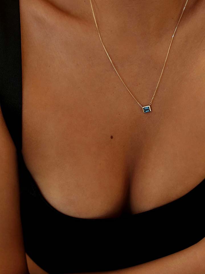 On Model shot of Necklace with thin yellow gold chain and blue rectangle topaz pendant. Model wearing black shirt and only her chest is in frame. 