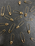 Product shot of yellow gold skull safety pins laid out on black stone with mini safety pins and regular yellow gold safety pins.  
