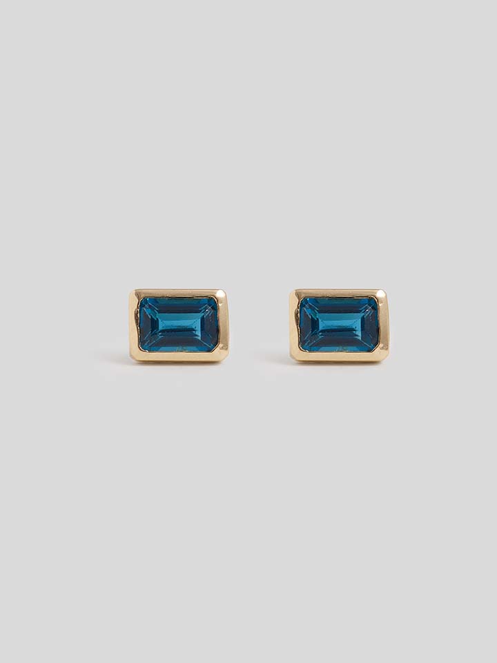 Product Shot of Vertical Blue Topaz Stud Earrings with gold post backing and gold trim around gem with white background 