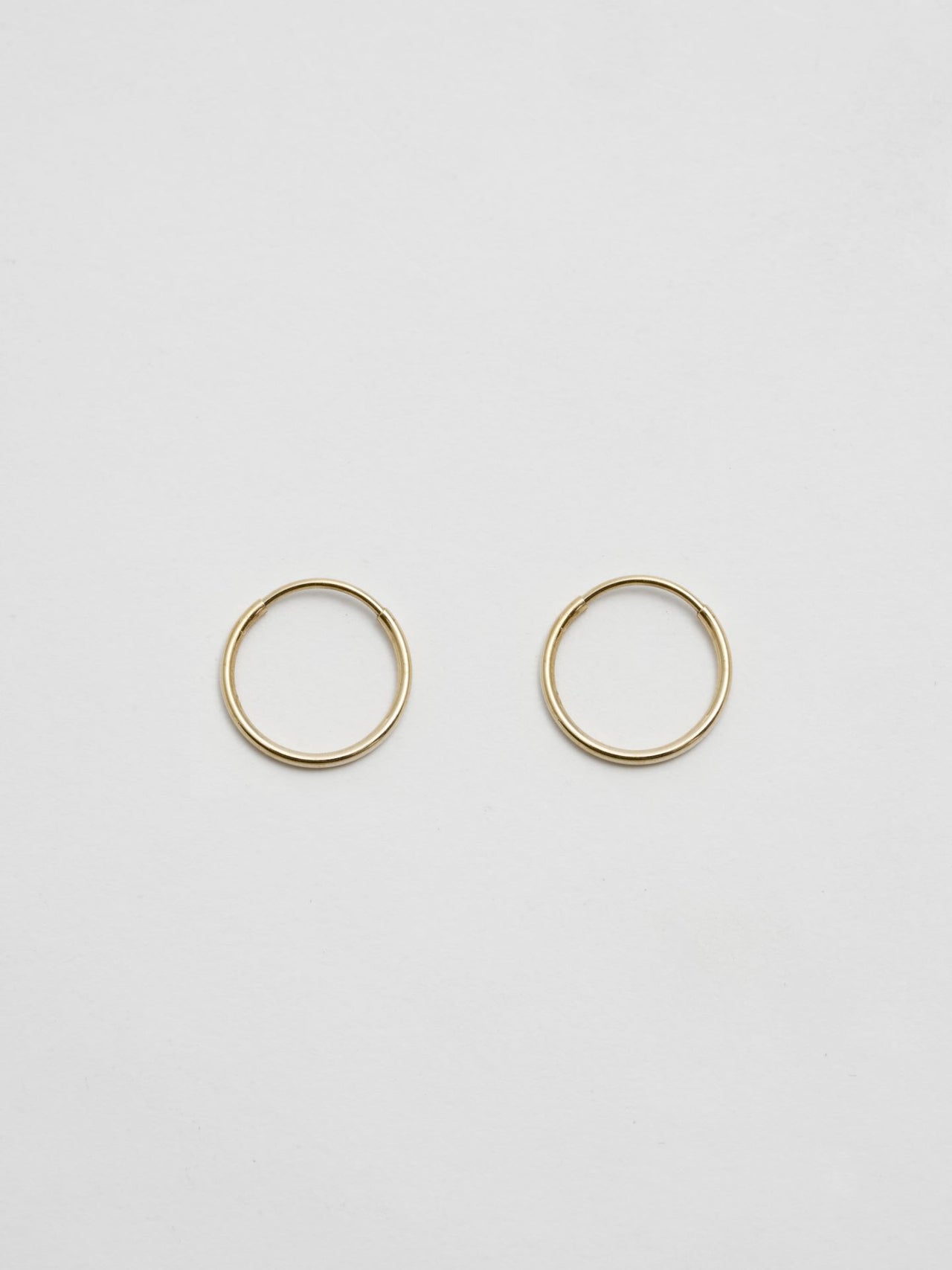 Front product shot of the Mini Infinity Hoops (14Kt shiny Yellow Gold Hoop Earrings  Diameter: 0.75”) Background: Grey backdrop