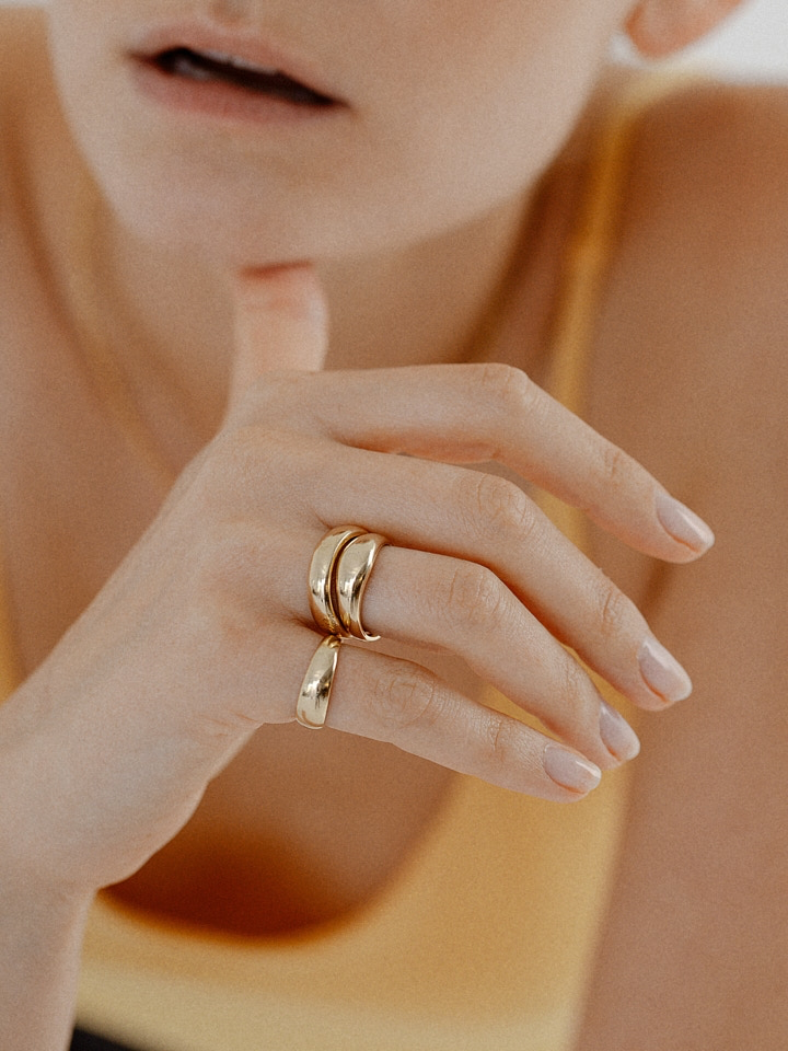 Close up shot of model’s hand wearing 2 Baby Dome Rings (shiny 14Kt Yellow Gold Dome Ring Tapered Width) stacked on ring finger, and 1 dome ring on pinky. Model's thumb placed on chin, background is model's chest out of focus in orange tank top. 