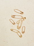 Flat Lay image of multiple Safety Pin Earring (14Kt Shiny Yellow Gold Safety Pin Earring Length: 21.35mm) Background: Beige fabric