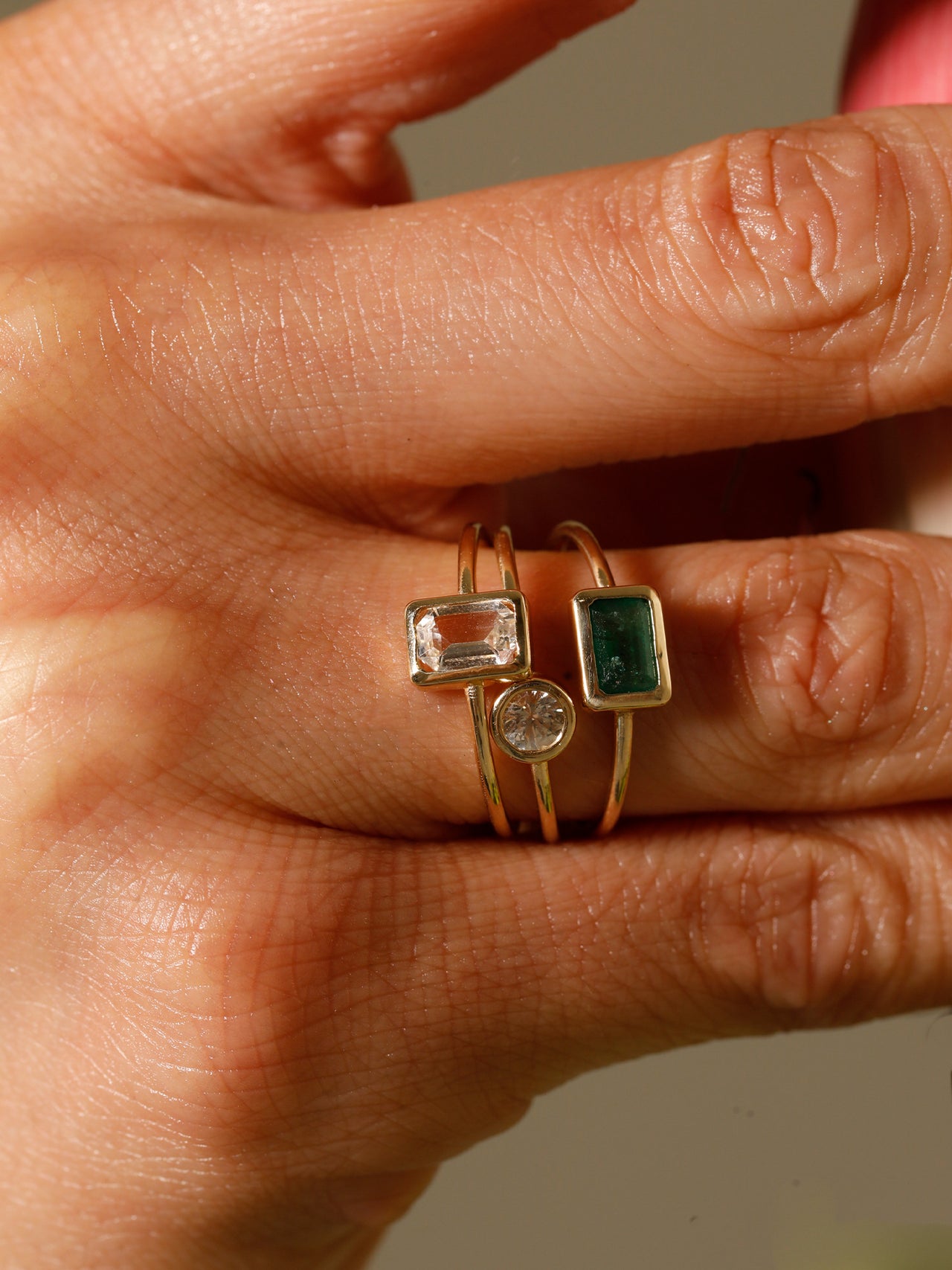 Close of of model’s fingers wearing an Emerald Cut Bezel Ring (Shiny Thin 14Kt Yellow Gold band with rectangular Green Quartz stone framed with yellow gold) stacked on top of thin yellow-gold banded ring with small round white topaz stone, and thin yellow-gold banded ring with small rectangular white topaz stone on ring finger. 